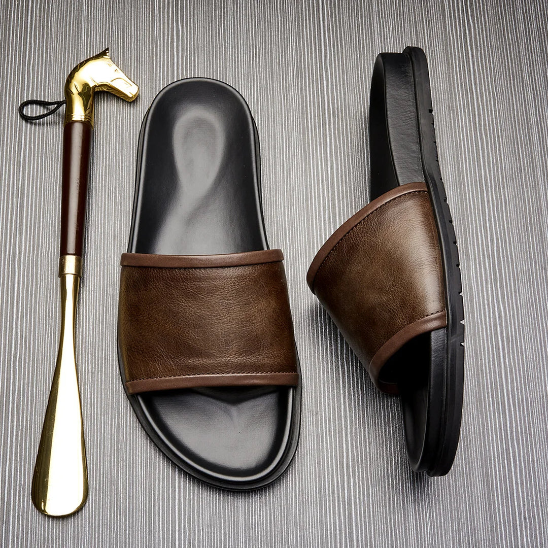 A pair of brown and black handmade leather sandals sit next to a gold shoehorn with a horse head handle, all placed on a textured gray surface.