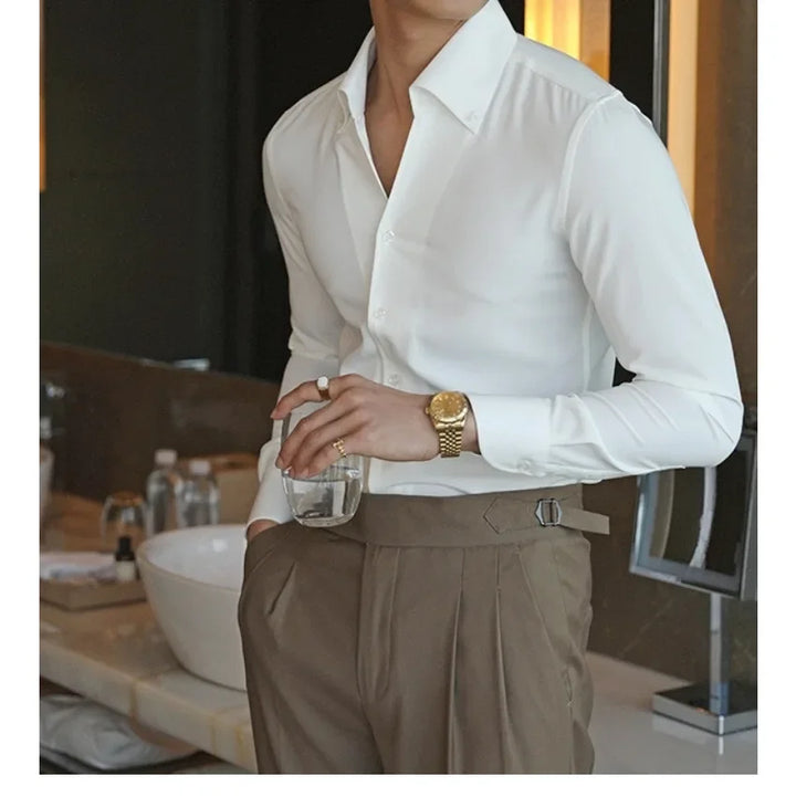 A person wearing a slim fit cotton bamboo shirt and brown trousers stands by a sink, holding a glass of water. Their face is not visible, and they have a gold watch on their left wrist.