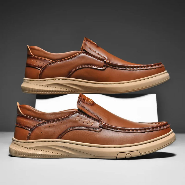 A pair of men's cowhide leather slip-on loafers with beige soft soles is shown side by side against a gray background. The shoes feature detailed stitching and perforated accents on the heel.