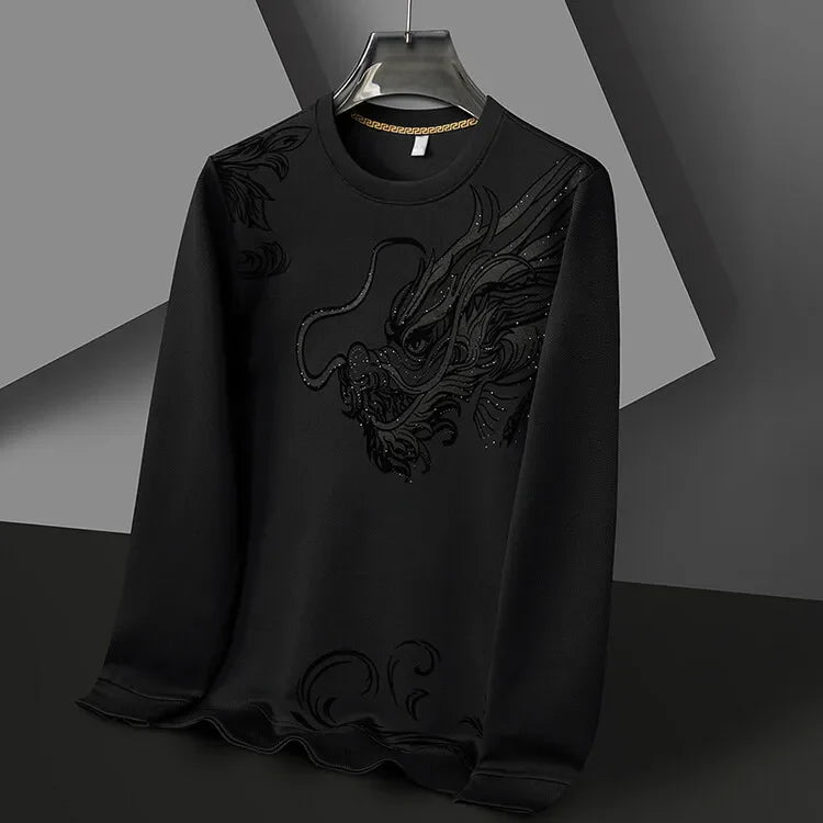 A black long-sleeve Year of the Dragon crewneck hangs on a hanger. It features a textured, embroidered dragon design on the front, blending subtly with ultra-soft cotton fabric. The background is composed of geometric shapes in grey and black.
