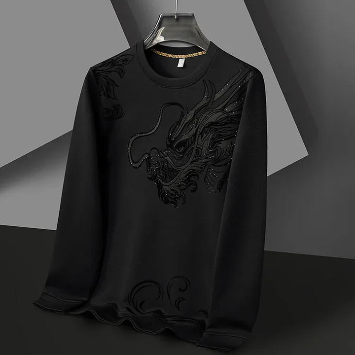 A black long-sleeve Year of the Dragon crewneck hangs on a hanger. It features a textured, embroidered dragon design on the front, blending subtly with ultra-soft cotton fabric. The background is composed of geometric shapes in grey and black.