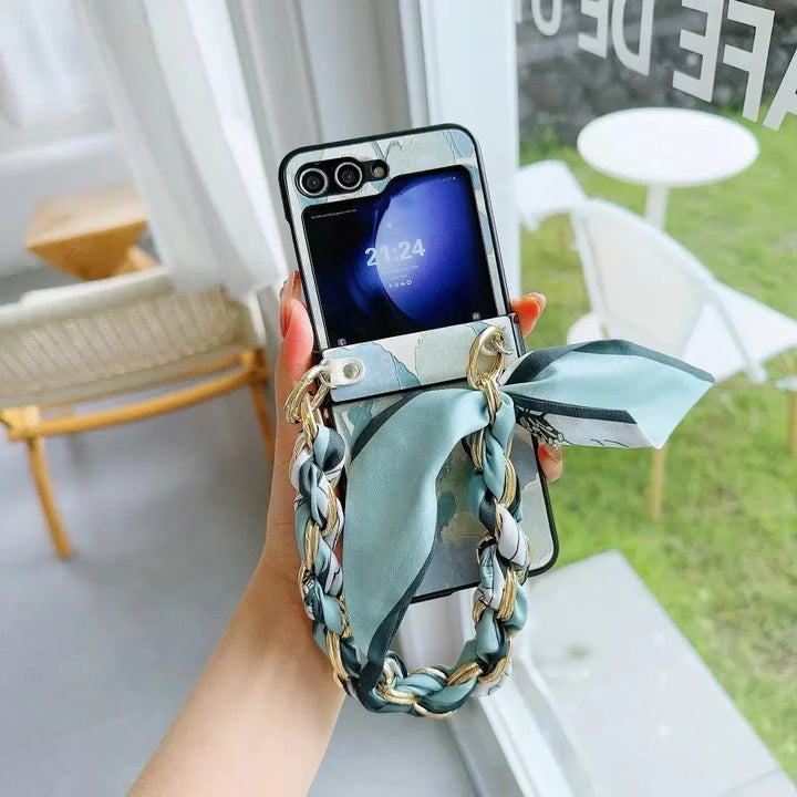 A hand holds a smartphone with a slim design and a stylish case featuring a blue-green chain strap and matching silk ribbon, in a bright room near a window overlooking a chair, table, and green grass outside.