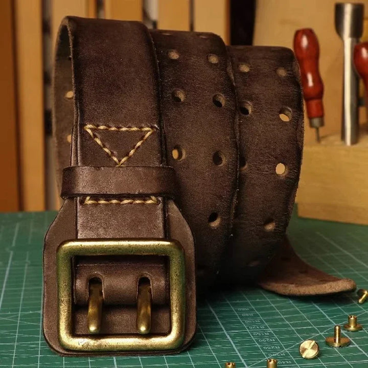 A handcrafted cowhide belt with a brass buckle is coiled on a green cutting mat, surrounded by leatherworking tools and rivets, embodying a rugged aesthetic.