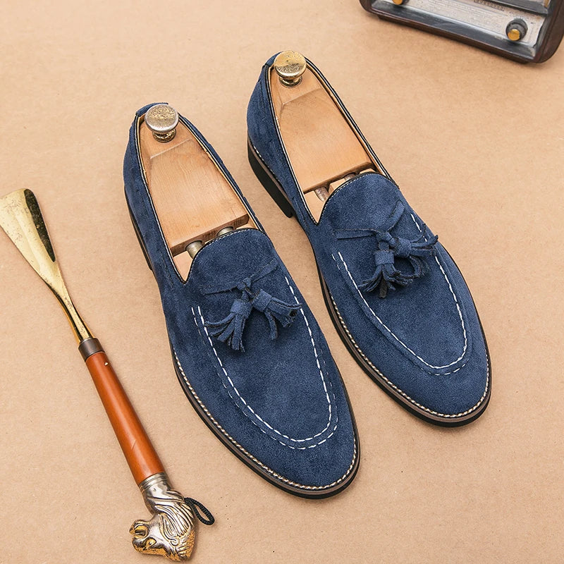 A pair of blue suede tassel loafers with white stitching, displayed with wooden shoe trees inside, on a tan surface next to a shoehorn with a lion head handle. Enjoy cushioned footbed comfort and durable construction in every step.