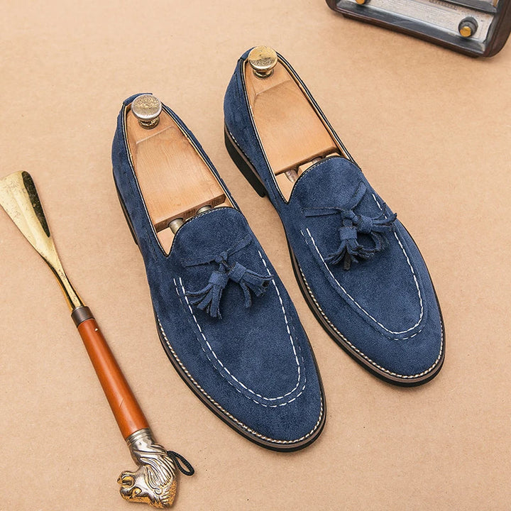 A pair of blue suede tassel loafers with white stitching, displayed with wooden shoe trees inside, on a tan surface next to a shoehorn with a lion head handle. Enjoy cushioned footbed comfort and durable construction in every step.