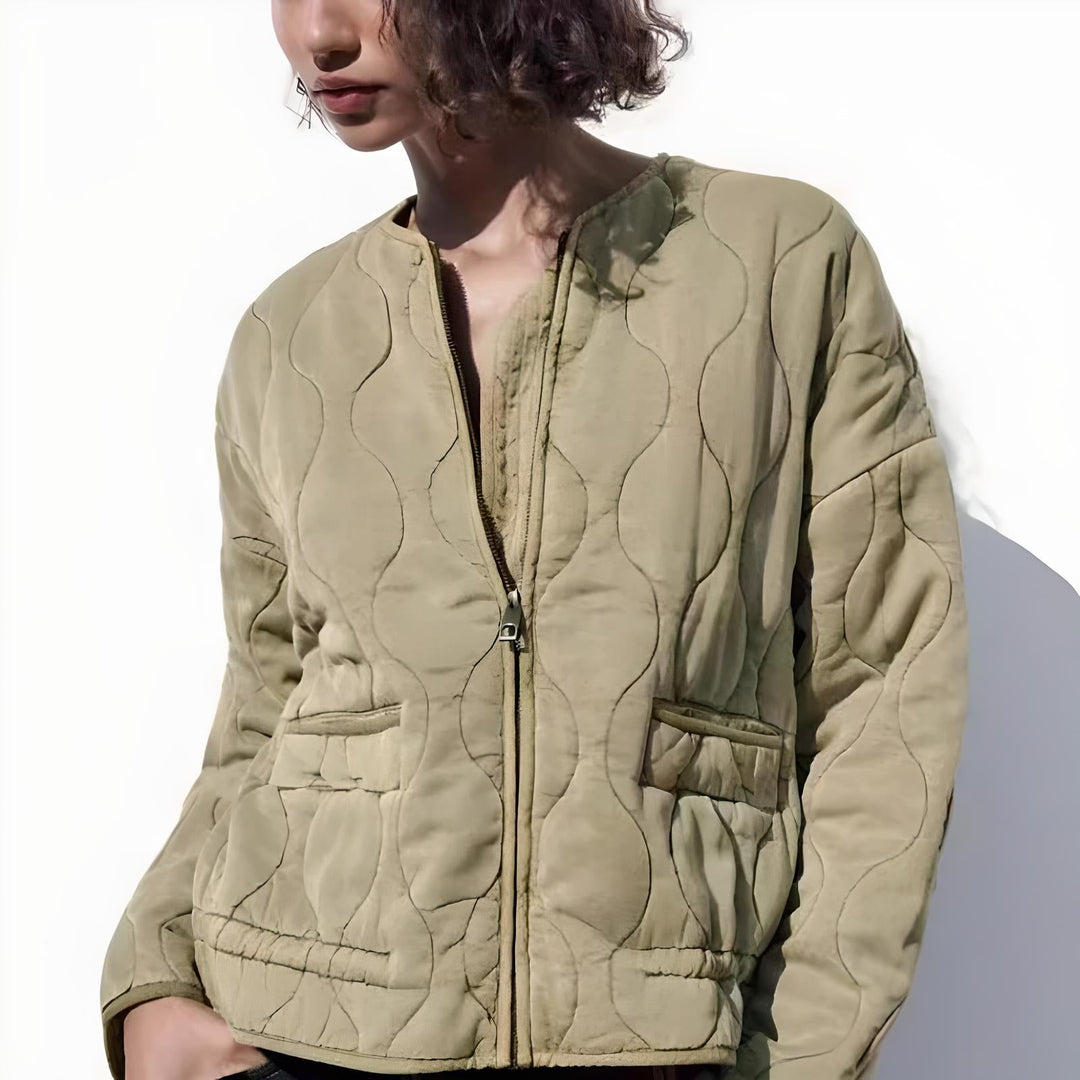 A person wears a light olive quilted bomber jacket with padded insulation, front zipper, and two pockets, standing against a plain white background. Only the upper body and lower half of the face are visible.