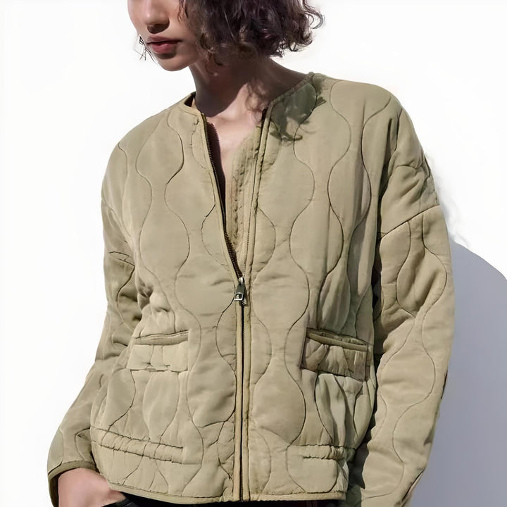 A person wears a light olive quilted bomber jacket with padded insulation, front zipper, and two pockets, standing against a plain white background. Only the upper body and lower half of the face are visible.