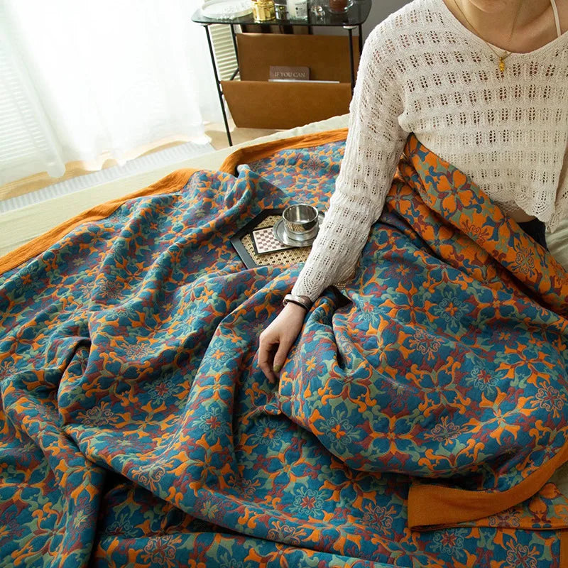 A person in a white sweater sits on a bed covered with a vibrant blue and orange ethnic pattern blanket, with a tray holding a teacup and book nearby in a sunlit room.