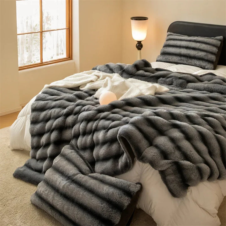 A cozy bedroom features a bed with a fluffy gray faux rabbit fur blanket and matching pillow, a white blanket, a glowing lamp, and natural light streaming through a window.