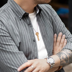 A person with folded arms wears a gray wrinkle-resistant pinstripe shirt over a white t-shirt, a silver watch, a tattoo on their forearm, and a key-shaped necklace. The persons face is partially visible.