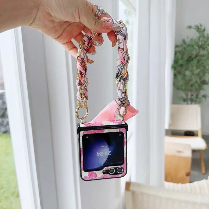 A hand holds a pink, floral-patterned flip phone with a decorative chain strap, in a bright room with large windows, a wooden chair, and a green plant in the background.