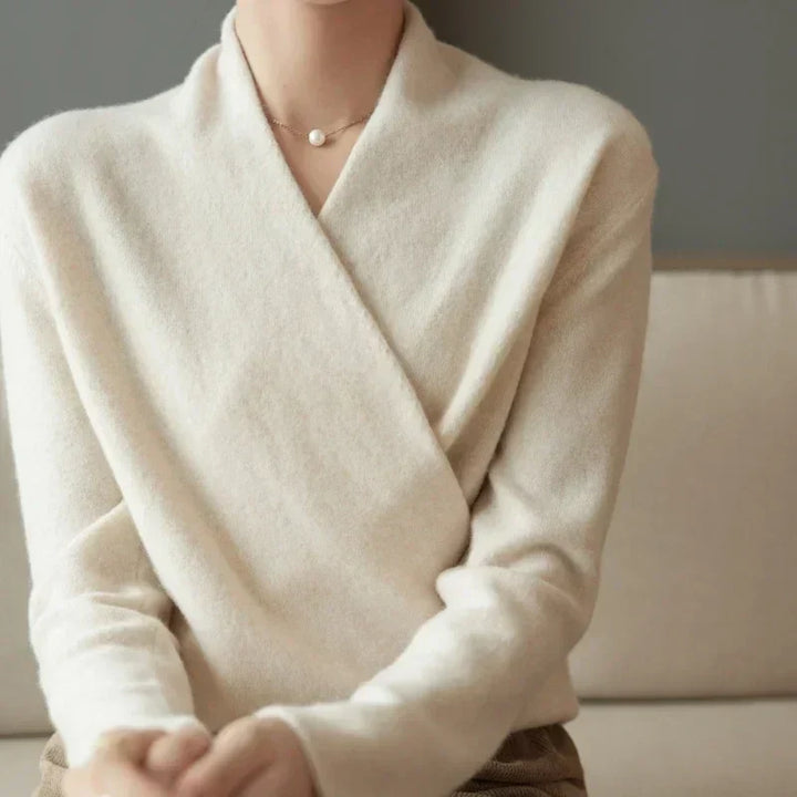 A person sits on a beige sofa wearing a soft, light beige cashmere wrap sweater and a delicate necklace with a small round pendant. The background is plain and the persons face is not fully visible.