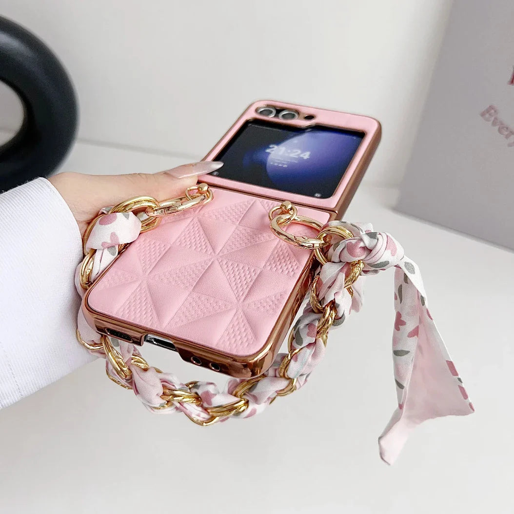 A hand holding a Samsung Galaxy Z Flip with a quilted premium leather case, featuring a decorative gold chain and a pink patterned scarf wrapped around the strap. The phones outer screen is visible.