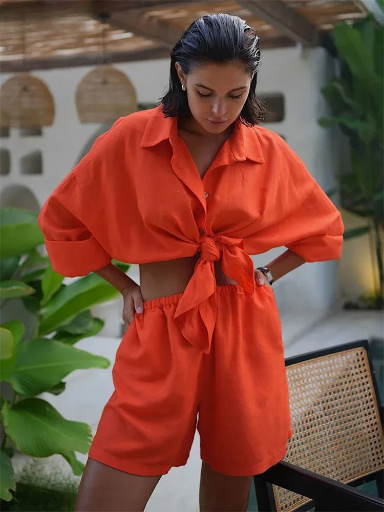 A woman stands outdoors in a bright orange 2 piece set—short-sleeved, button-up shirt tied at the waist and matching high-waisted shorts. She looks down, surrounded by lush green plants and modern decor.