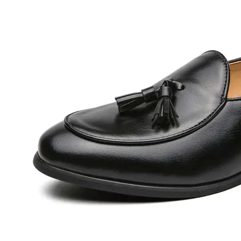 A close-up of a black leather loafer with tassels on the upper, featuring a vintage look, shows the front and side of the shoe against a white background.
