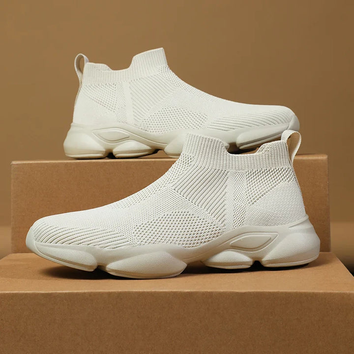 A pair of white slip-on athletic sneakers with breathable knit fabric uppers and chunky non-slip soles, displayed on stacked brown cardboard boxes against a brown background.