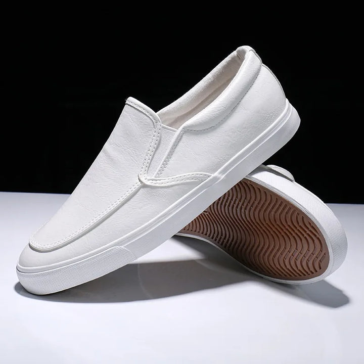 A pair of mens slip-on white sneakers with rubber soles, one shoe resting on its side and the other upright, showcasing the brown, textured outsole against a black and white background.