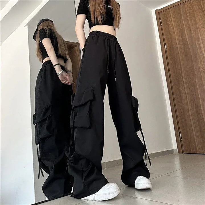 A person in loose black cargo pants with an adjustable drawstring waist and cargo pockets, a cropped black top, and white sneakers stands indoors, their reflection visible in the mirror behind them.
