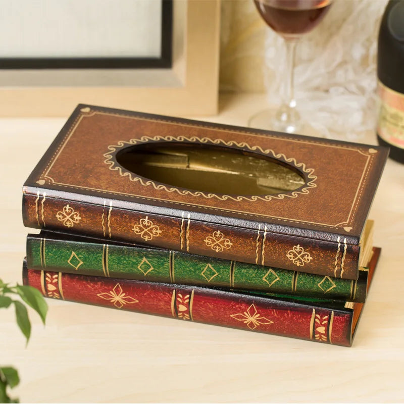 A decorative tissue box cover designed to look like two stacked vintage books sits on a light wooden surface, with a glass of red wine and a plant in the background—perfect for home library decor.