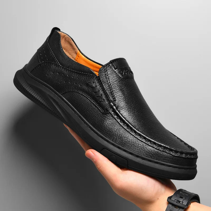 A person holds a black Men's Cowhide Leather Shoe with visible stitching, soft sole, and slip-on design against a plain light gray background.