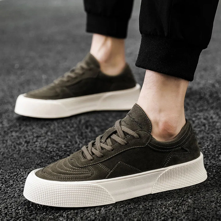 A person wearing dark green suede lace-up sneakers with thick white rubber soles, standing on a textured black surface. The comfortable cushioning of these shoes pairs well with their black pants.
