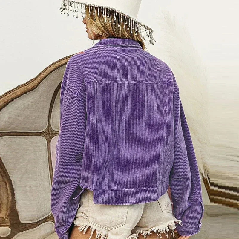 A person wearing a purple corduroy jacket with sequin floral patches and frayed white denim shorts stands with their back to the camera, also wearing a cream wide-brimmed hat with dangling beads.