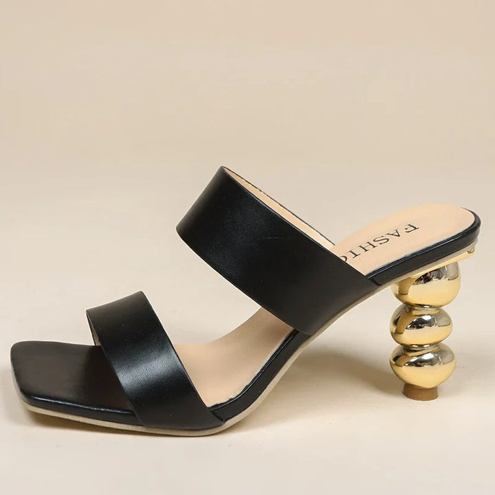 A black high-heeled sandal crafted from premium leather with two wide straps and a unique sculpted gold heel made of stacked, round metallic shapes. The cushioned footbed is beige and the toe is square-shaped.