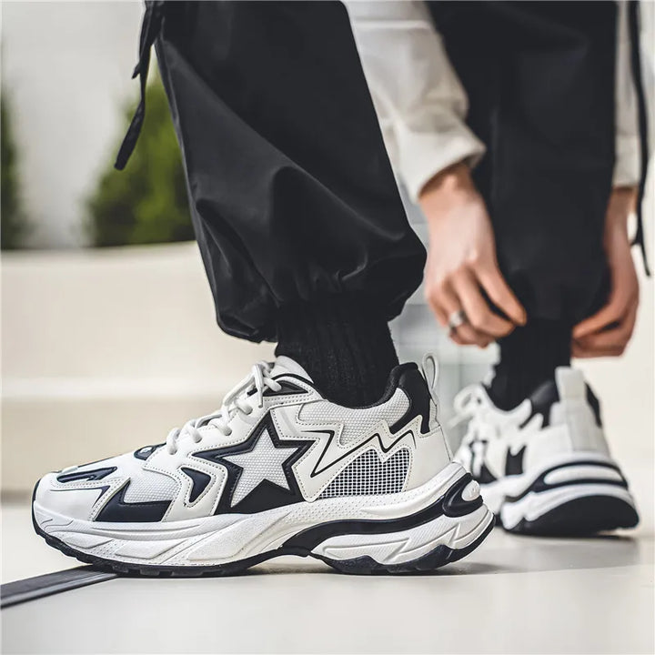 A person wearing black pants and socks is lacing up stylish black and white sneakers with star designs, featuring a breathable mesh upper, while standing on a light-colored floor.