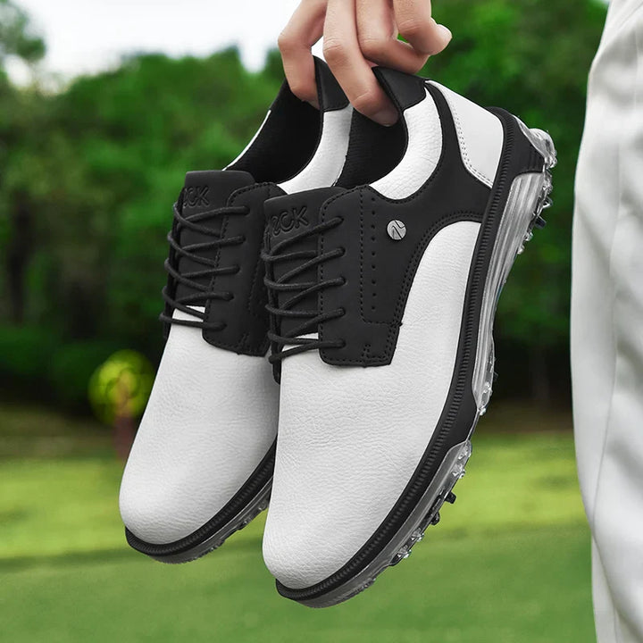 A person holds a pair of white and black athletic shoes with black laces and transparent rubber soles outdoors, featuring advanced grip technology, with green grass and trees in the blurred background.