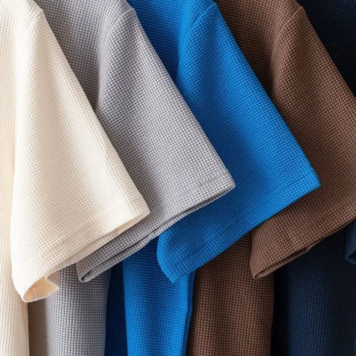 Five short-sleeved, waffle-textured shirts made from high-quality cotton in cream, gray, blue, brown, and dark navy are hanging side by side, partially overlapping each other.