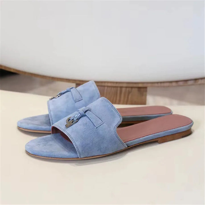 A pair of light blue slide sandals with a suede finish, flat soles, open toes, and decorative buckles on the upper straps, placed on a light surface.