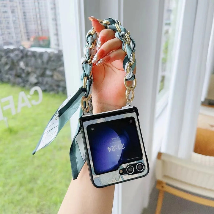 A hand holds a flip smartphone with a slim design and decorative chain strap, plus a patterned scarf attached, in front of large windows overlooking buildings and greenery.