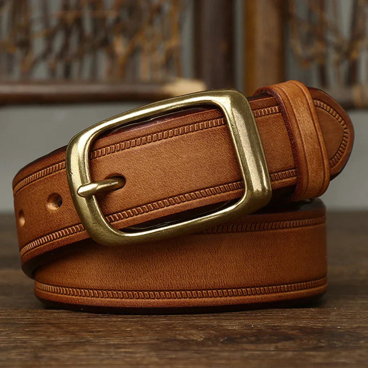 A tan cowhide belt with detailed stitching and a large, rectangular brass buckle is coiled and set on a wooden surface with a blurred background.