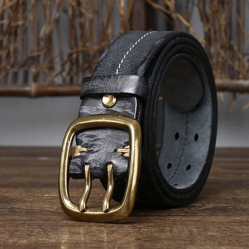 A rolled-up genuine leather belt with white stitching and a gold-colored double prong buckle sits on a wooden surface, showcasing heritage craftsmanship against a rustic background.