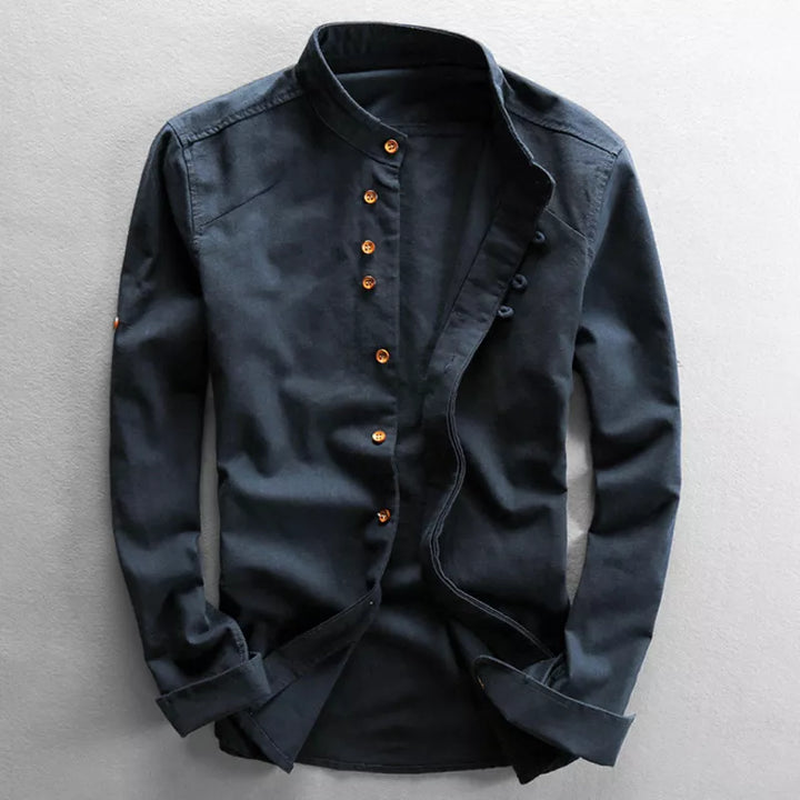 A dark blue, long-sleeved, button-up shirt with a mandarin collar. This Japanese inspired overshirt features distinctive brown buttons arranged slightly off-center and an open front, displayed against a light gray background.