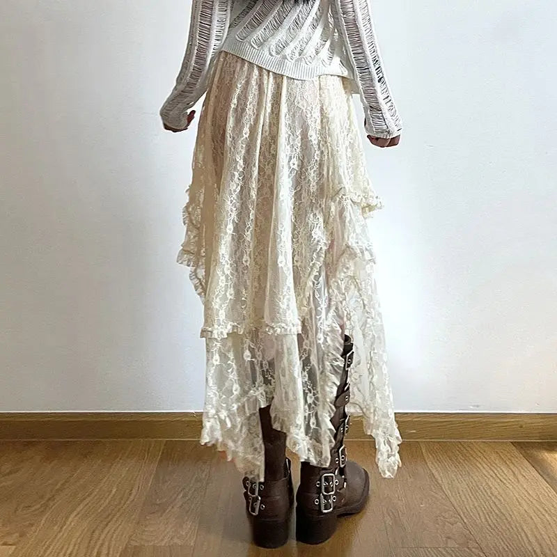 A person stands on a wooden floor wearing a white, high waist asymmetrical hemline lace skirt, a white long-sleeve top, and brown buckled boots, facing away from the camera against a plain white wall.