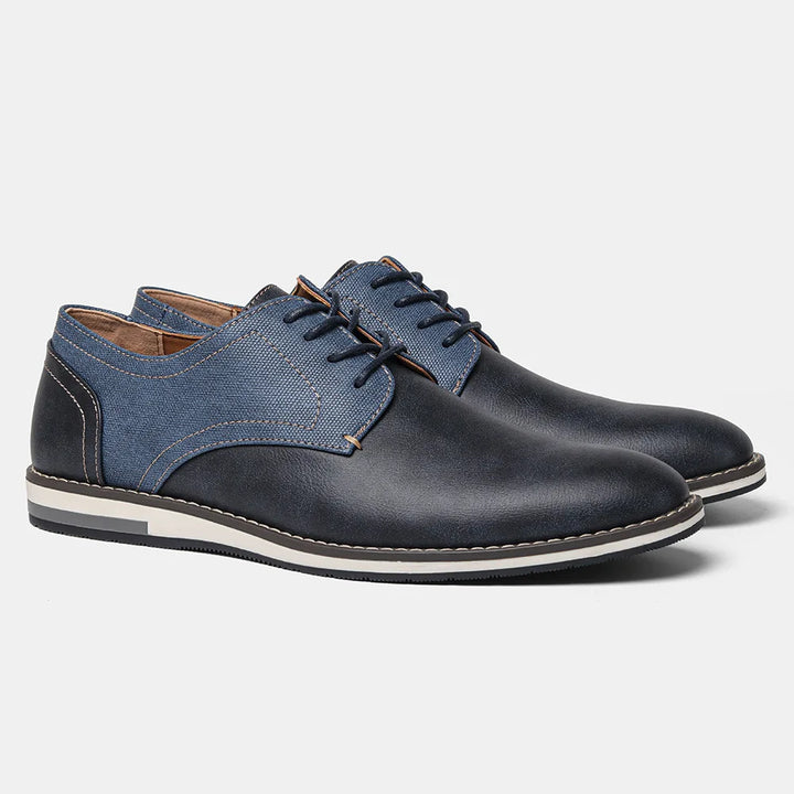 A pair of stylish patchwork oxfords for men, featuring dark blue leather and denim-like panels, white stitching, contrasting white and black rubber soles, and a cushioned sole for all-day comfort.