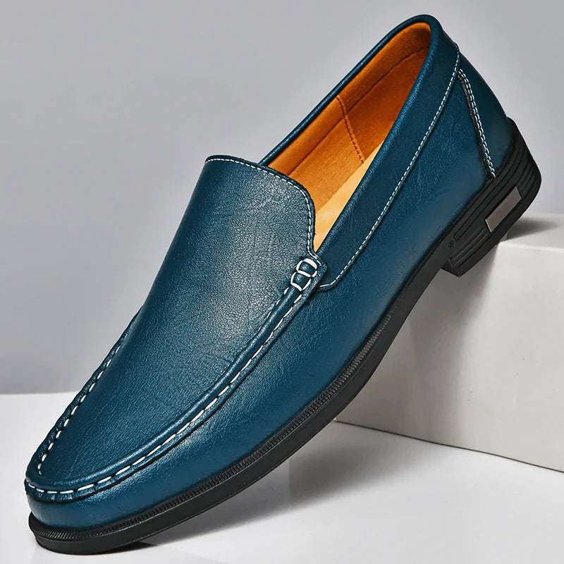 A single teal blue classic slip-on loafer in genuine leather, featuring white stitching, a black sole, and a tan inner lining, is displayed on a white surface, slightly elevated at the back.