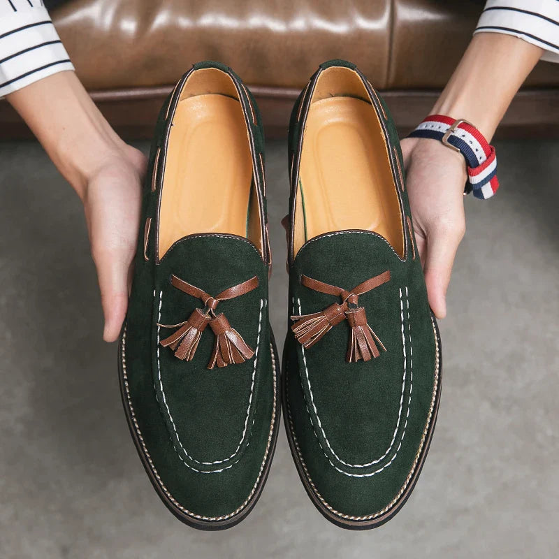 A person holds a pair of dark green suede tassel loafers with brown tassels and white stitching, showcasing the shoes and their durable construction above a gray floor.