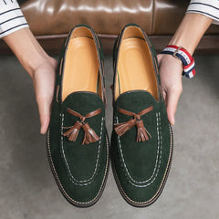 A person holds a pair of dark green suede tassel loafers with brown tassels and white stitching, showcasing the shoes and their durable construction above a gray floor.