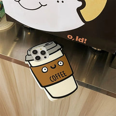 A phone in a 3D coffee cup case with a smiling face and COFFEE printed on it, made of soft silicone for shockproof protection, rests on a wooden counter next to a sign with part of a cartoon face.