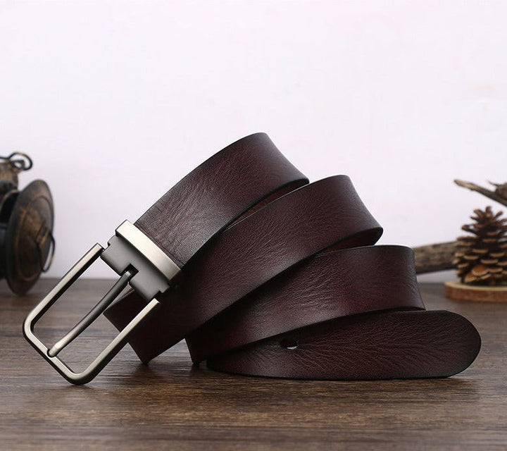 A dark brown cowhide belt with a silver buckle is coiled on a wooden surface. In the background, there is a pine cone and some decorative items, all against a light wall.