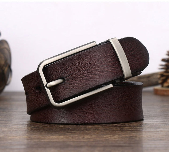 A dark brown cowhide belt with a silver rectangular buckle, coiled neatly on a wooden surface with blurred background decor.
