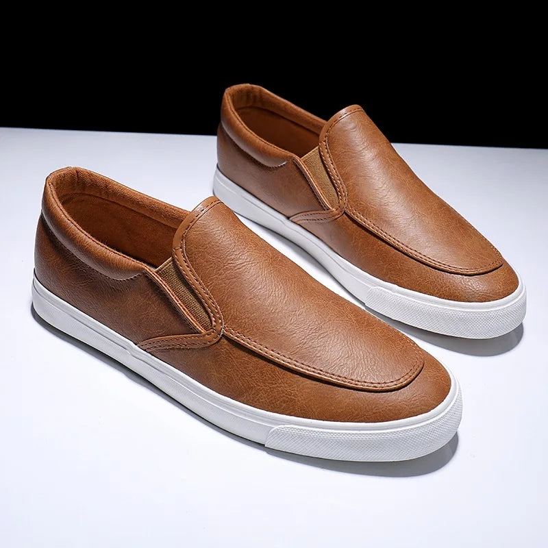 A pair of brown mens slip-on casual shoes with white rubber soles and a comfortable leather insole, displayed on a white surface against a dark background.