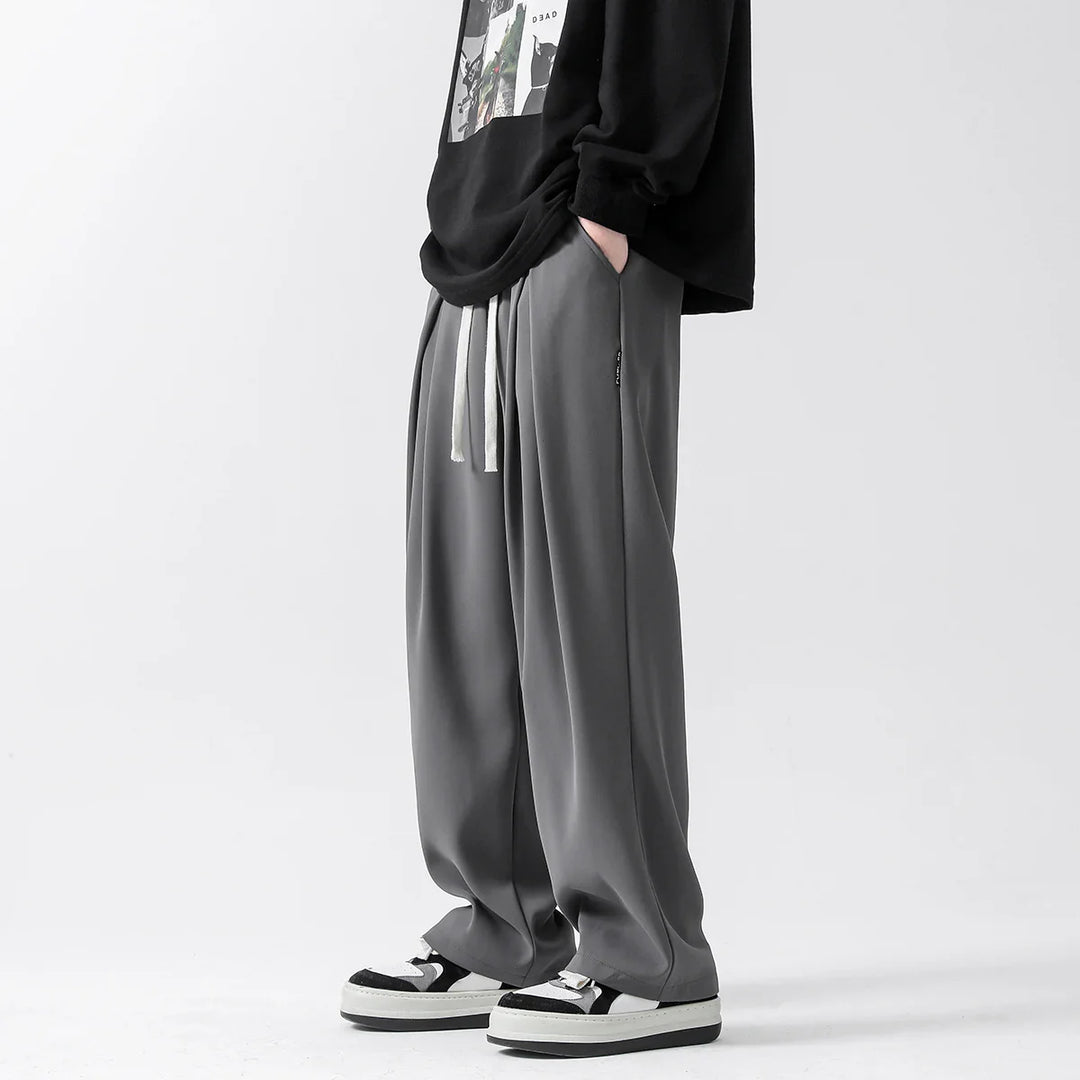 A person stands with hands in pockets, wearing loose-fitting gray streetwear joggers with white drawstrings, a black graphic sweatshirt, and black-and-white sneakers against a plain light background.