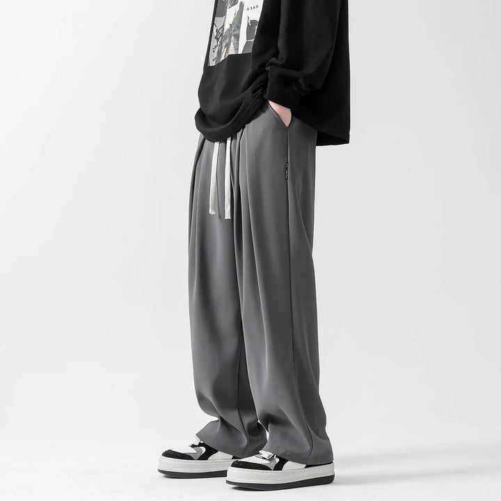 A person stands with hands in pockets, wearing loose-fitting gray streetwear joggers with white drawstrings, a black graphic sweatshirt, and black-and-white sneakers against a plain light background.