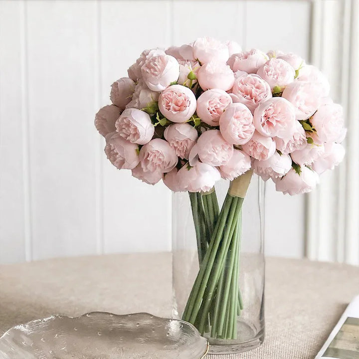 A bouquet of maintenance-free pale pink peonies with green stems arranged in a clear glass vase, placed on a round beige table with a clear decorative plate nearby against a light-colored background.
