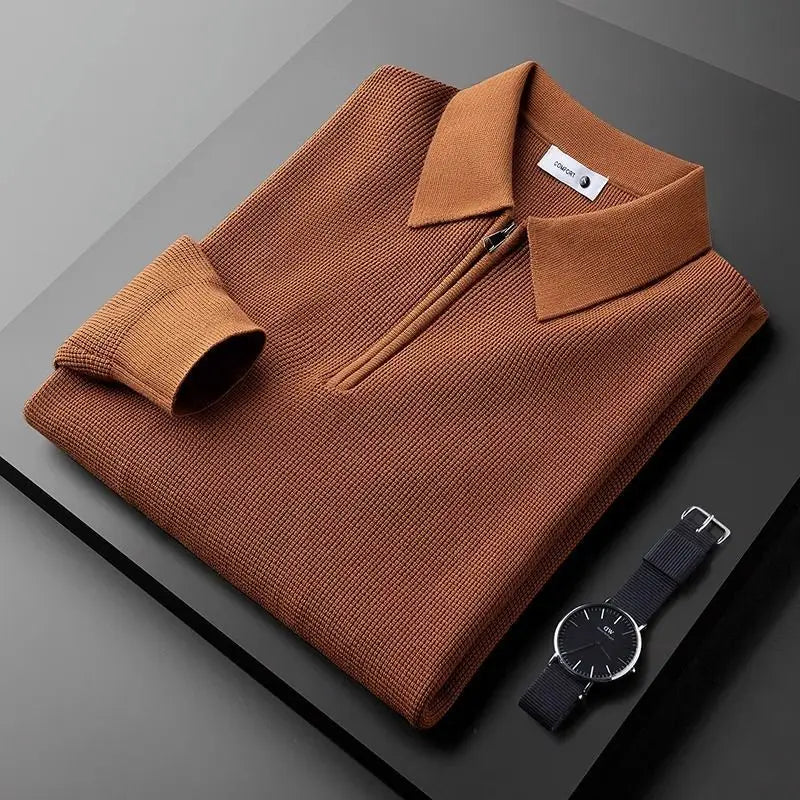 A neatly folded brown zip-collar sweater made from stretchy fabric is displayed on a dark surface next to a black wristwatch with silver details. The sweater’s sleeve is folded once, and a tag is visible inside the collar.