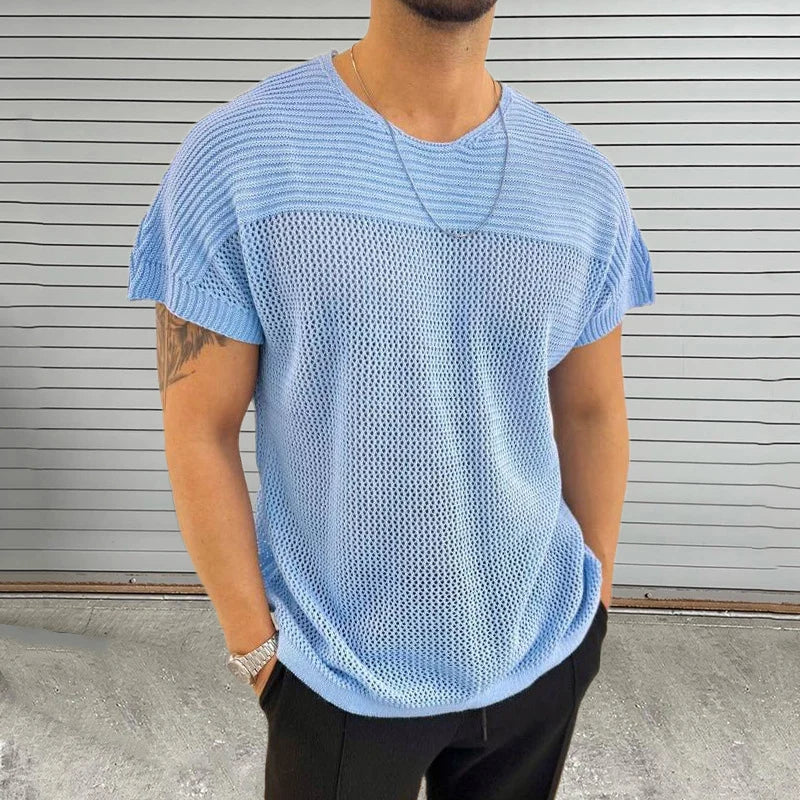 A person wearing a light blue, short-sleeved mesh knit tee and black pants stands in front of a gray, horizontal-striped metal wall. The breathable design pairs with streetwear accessories—a tattoo, thin necklace, and watch—for a modern look.