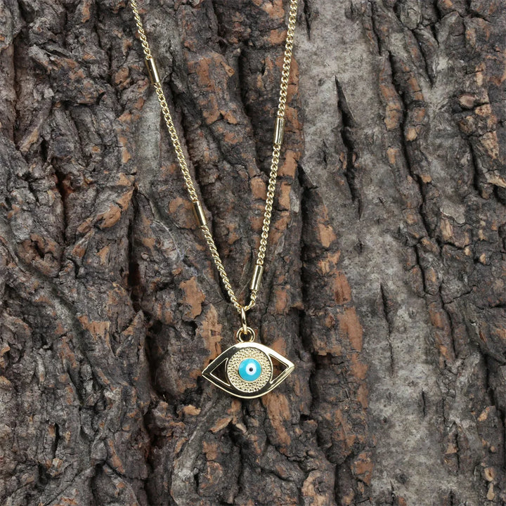 A hypoallergenic gold evil eye necklace with a blue zirconia stone pendant is displayed against the rough, textured bark of a tree.